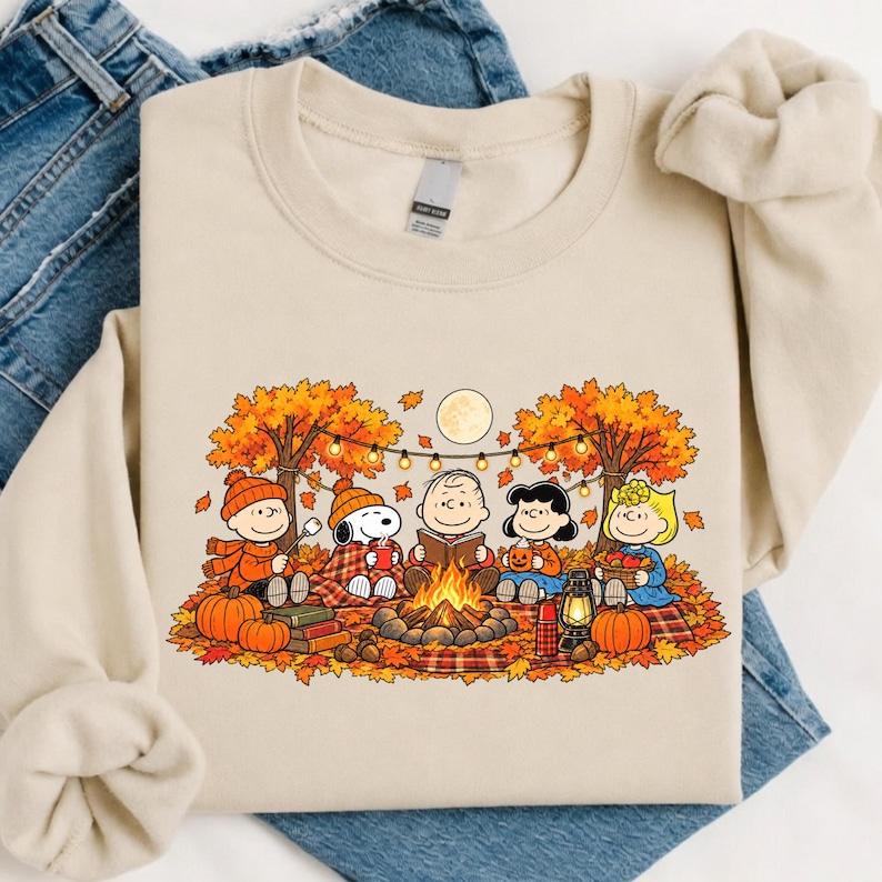 May include: A beige crewneck sweatshirt featuring a colorful graphic of the Peanuts characters sitting around a campfire in an autumn forest. The scene includes orange fall leaves, pumpkins, a string of lights, and a full moon in the background.