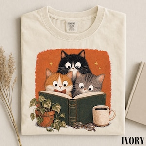 May include: An ivory t-shirt featuring a graphic of three cats reading an open green book. The illustration includes a potted plant, a coffee mug, and round glasses. The cats are black, orange, and grey. The background is a textured orange square.