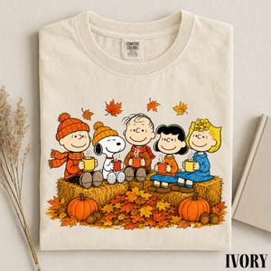 May include: An ivory t-shirt featuring a graphic of the Peanuts characters Charlie Brown, Snoopy, Linus, Lucy, and Sally sitting on hay bales. They are wearing autumn clothing, holding mugs, and surrounded by falling orange and yellow leaves.