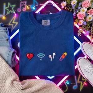 Puede incluir: Una camiseta azul Comfort Colors con gráficos de corazón pixelado, Wi-Fi, auriculares y monopatín. La camiseta está rodeada de una luz de neón, flores y un par de zapatillas blancas. Unos vaqueros y un jersey también están en la imagen.