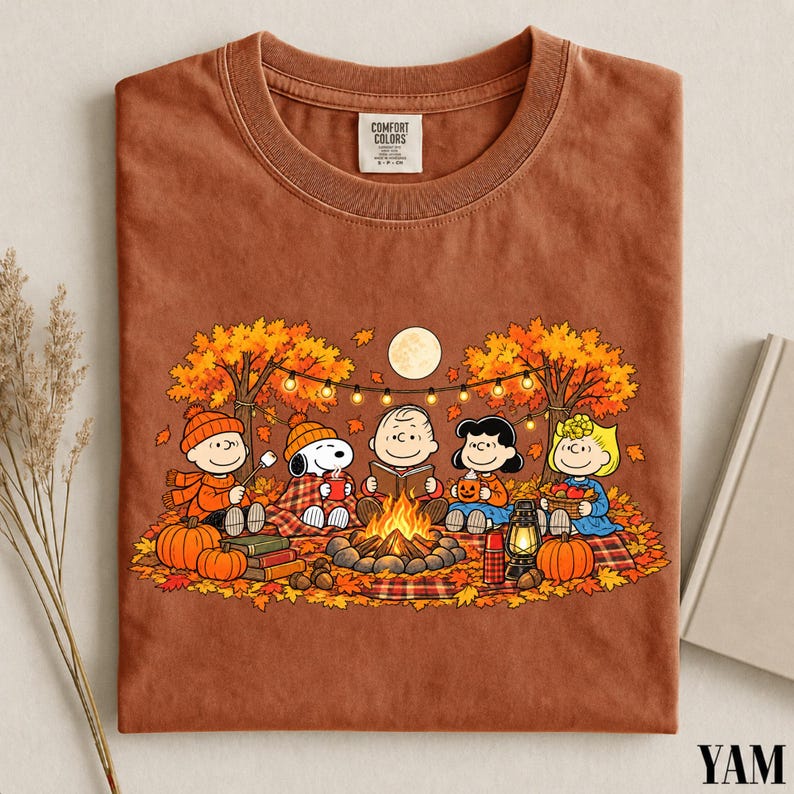 May include: A rust-colored t-shirt with a graphic print featuring the Peanuts characters sitting around a campfire in an autumn forest. The design includes Snoopy, Charlie Brown, and friends with pumpkins, a lantern, and string lights. The neck label reads Comfort Colors.