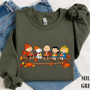 May include: An olive green crewneck sweatshirt featuring a graphic of the Peanuts characters sitting on a wooden fence, each holding a mug. The design includes Snoopy, Charlie Brown, and friends wearing autumn hats and scarves with orange pumpkins.