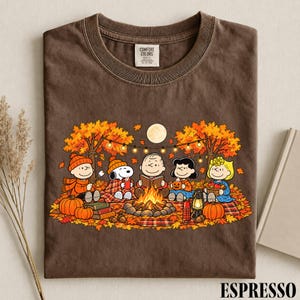 May include: A brown crew neck t-shirt featuring a graphic print of the Peanuts characters sitting around a campfire in an autumn forest. The design includes Snoopy, Charlie Brown, and friends with pumpkins, a lantern, and string lights.