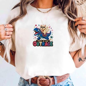 Puede incluir: Camiseta color crema con un gráfico colorido de un gato con gafas de sol montado en un dinosaurio azul. El texto "THIS IS AN OUTRAGE" está debajo en un degradado arcoíris.