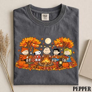 May include: A charcoal gray crew neck t-shirt featuring a graphic print of the Peanuts characters sitting around a campfire in an autumn forest. The design includes Snoopy, Charlie Brown, and friends with pumpkins, a lantern, and string lights.