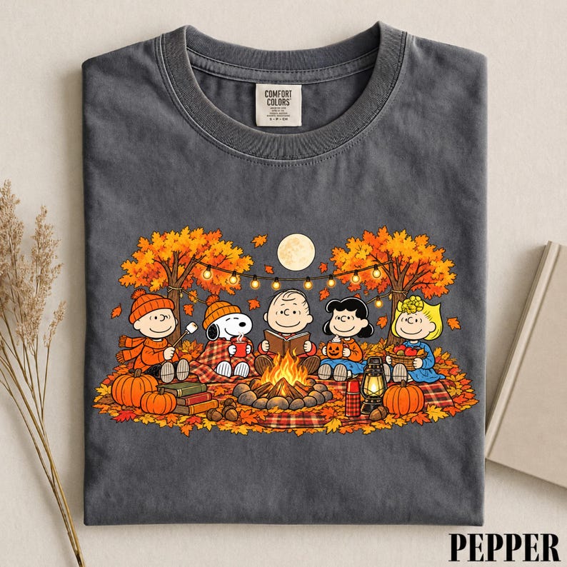 May include: A charcoal gray crew neck t-shirt featuring a graphic print of the Peanuts characters sitting around a campfire in an autumn forest. The design includes Snoopy, Charlie Brown, and friends with pumpkins, a lantern, and string lights.