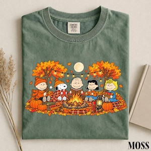 May include: A moss green crew neck t-shirt featuring a graphic of the Peanuts characters sitting around a campfire in an autumn forest. The design includes Snoopy, Charlie Brown, and friends with pumpkins, a lantern, and string lights under a full moon.