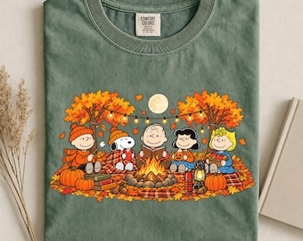 May include: A moss green crew neck t-shirt featuring a graphic of the Peanuts characters sitting around a campfire in an autumn forest. The design includes Snoopy, Charlie Brown, and friends with pumpkins, a lantern, and string lights under a full moon.