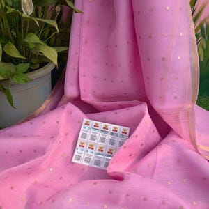 May include: A draped pink fabric with gold square accents. The fabric has a subtle sheen and is folded to reveal its texture. A small sticker sheet with QR codes is visible on the fabric. A potted plant is in the background.