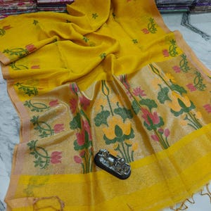 May include: A yellow textile with floral patterns in green, pink, and yellow. The fabric has a shimmering quality and a fringed edge. The design includes stylized flowers and leaves.