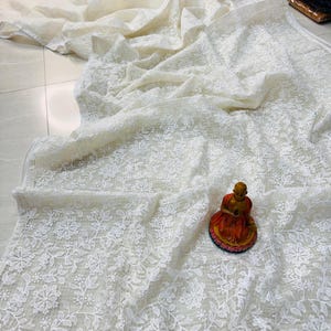 Könnte beinhalten: Ein transparenter, cremeweißer Stoff mit aufwendigen weißen Blumenstickereien auf der gesamten Fläche. Das Textil weist eine subtile gitterartige Struktur auf und ist in weichen Falten drapiert, begleitet von einer kleinen sitzenden Figur.