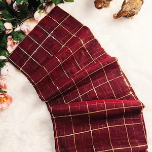 May include: A deep maroon fabric piece featuring a gold grid pattern and a row of matching maroon tassels along the bottom edge. The textile has a textured appearance and is arranged in folded layers, showcasing the consistent square design.