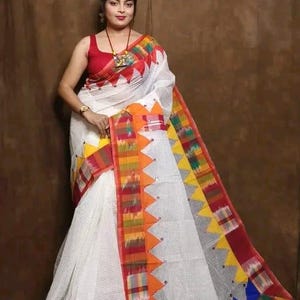 May include: A white saree featuring a grid pattern and a vibrant border with multicoloured geometric triangles in red, orange, yellow, green, and blue. The fabric includes decorative mirror accents along the edges and a red sleeveless blouse.