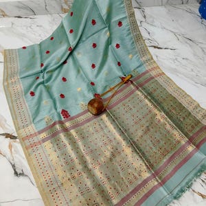 Könnte beinhalten: Ein hellblauer Sari aus Seide mit kleinen roten Blumenmotiven und goldfarbenen Akzenten. Der Stoff hat einen breiten, verzierten Rand mit komplexen goldenen Metallmustern und dünnen roten Streifen. Ein kleiner Holzgegenstand liegt auf dem drapierten Stoff.