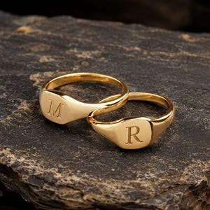 May include: Two gold signet rings with polished surfaces, each featuring a single engraved letter. One ring is engraved with the letter M, and the other ring is engraved with the letter R. The rings have a smooth, rounded band and a flat face.