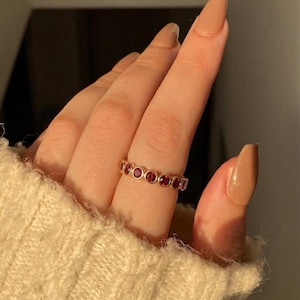 Puede incluir: Un anillo de banda en tono dorado que presenta una hilera continua de pequeñas piedras preciosas redondas de color rojo intenso, engastadas en biseles circulares individuales. El anillo se lleva en un dedo, mostrando la disposición uniforme de las piedras.