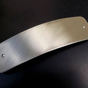 May include: A silver-colored metal bar with a brushed finish and rounded edges. The bar has two small, round holes on either end.