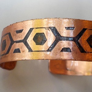 May include: Copper cuff bracelet with a geometric pattern. The bracelet has a series of black hexagon and diamond shapes etched into the copper. The cuff is open and has a slightly textured surface.