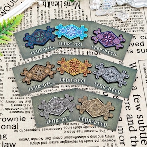 Może przedstawiać: Kolekcja ozdobnych przypinek z geometrycznymi wzorami w różnych kolorach, w tym niebieskim, złotym i srebrnym. Każda przypinka oznaczona jest napisem "LEOR-DECO".