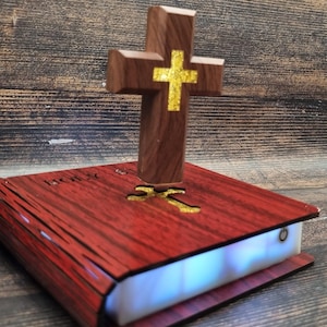 Peut inclure: Une croix en bois avec un centre doré est posée sur un objet rouge en forme de livre. Le livre porte le mot "HOLY" sur la couverture et émet une douce lumière bleue de ses pages. La croix est en bois foncé.