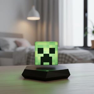 May include: A green, pixelated cube lamp with a Creeper face design, levitating above a black base. The lamp is illuminated, casting a soft green glow. The background shows a bedroom setting with a bed and a window.