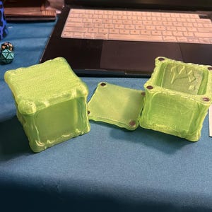 May include: Three lime green, 3D-printed boxes with textured surfaces. One box is closed, one is open, and a lid is separate. The boxes are on a blue surface, with a laptop and dice visible in the background. The boxes are likely for storage.