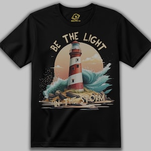 Könnte beinhalten: Ein schwarzes T-Shirt mit kurzen Ärmeln und einer Grafik eines rot-weiß gestreiften Leuchtturms auf einer felsigen Insel mit blauen Wellen und einem Sonnenuntergang. Der Text "BE THE LIGHT" steht oben und "IN THE STORM" unten.