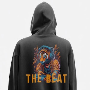 Op de afbeelding: Een antracietgrijze hoodie met een afbeelding op de achterkant van een tijger met een blauwe koptelefoon en een oranje jas, terwijl deze een muziekpaneel bedient. De tekst THE BEAT staat in grote oranje hoofdletters onder de tijgerafbeelding.