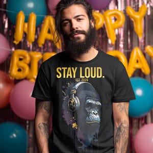Könnte beinhalten: Ein schwarzes Kurzarm-T-Shirt mit einer Grafik eines Gorillas, der Kopfhörer trägt. Der Text "STAY LOUD." steht in Gelb über dem Gorilla, darunter "EST. 2026". Die Grafik enthält violette und gelbe Farbspritzer-Effekte im Hintergrund.