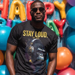 Könnte beinhalten: Schwarzes T-Shirt mit goldenem Text "STAY LOUD. EST. 2026". Das Shirt zeigt eine Grafik eines Gorillas mit Kopfhörern. Der Hintergrund umfasst bunte Luftballons und das Wort "HAPPY" in großen aufblasbaren Buchstaben.