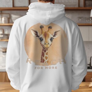 Op de afbeelding: Een wit sweatshirt met capuchon en een afbeelding op de achterkant van een giraffekop en -hals in een beige cirkel. Onder de giraffe staat de tekst REACH FOR MORE in een beige, schreefloos lettertype, gecentreerd op de stof.