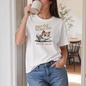 Puede incluir: Una camiseta blanca de manga corta con un gráfico de un gato atigrado marrón con el texto Paws Out Claws Out y You're On Thin Ice. El gato aparece en posición agachada con una expresión decidida, representado en un estilo de dibujos animados.