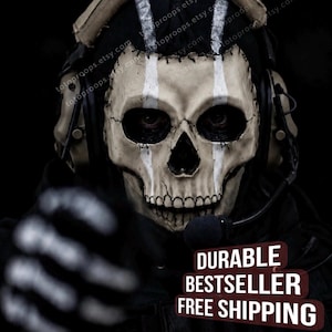 Könnte beinhalten: Eine taktische Totenkopfmaske mit beigefarbener Knochenoptik, schwarzen Augenhöhlen und vertikalen weißen Streifen. Die Maske wird mit einem beigen taktischen Headset und einem Mikrofon getragen. Der Text lautet DURABLE BESTSELLER FREE SHIPPING.