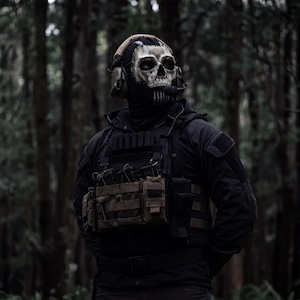 May include: A person wearing a skull mask, tactical vest, and headset stands in a dark forest. The mask is white with black details, and the vest has multiple pouches. The background features tall, dark trees.