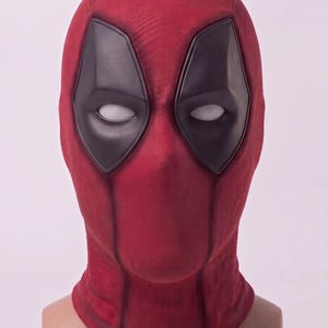 Può includere: Una maschera integrale in tessuto rosso scuro testurizzato, caratterizzata da ampie zone oculari nere e lenti a rete bianche. La maschera copre interamente la testa e il collo, presentando un design strutturato con cuciture e contorni visibili.