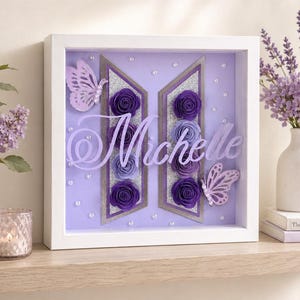 May include: A square white shadow box frame containing a purple background with a silver-bordered logo shape filled with purple paper roses. The name Michelle is written in white script across the center, accented by two light purple butterflies.
