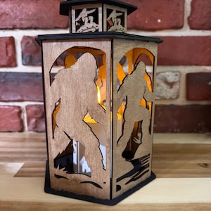 May include: A hexagonal wooden lantern featuring laser-cut silhouettes of Bigfoot figures on each panel. The lantern has a black top with a circular handle and a black base. A warm light glows from within, illuminating the cutouts of the creatures.