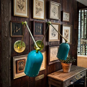 Peut inclure: Deux lampes murales turquoise avec des accents en laiton sont présentées. Elles sont fixées sur un mur en lambris de bois sombre, orné d'estampes botaniques encadrées et d'œuvres d'art vintage. Une petite table en bois et une plante en pot sont également visibles.