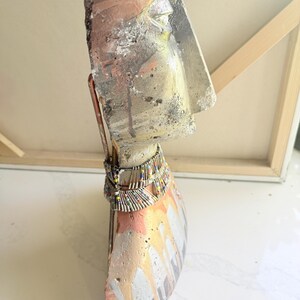 K&ouml;nnte beinhalten: Abstrakte Skulptur eines Kopfes und eines Torsos mit strukturierter, verwitterter Optik. Die Skulptur hat eine Farbpalette aus Silber, Orange und Wei&szlig; mit einer Perlenkette. Das Kunstwerk wird auf einer wei&szlig;en Oberfl&auml;che pr&auml;sentiert.