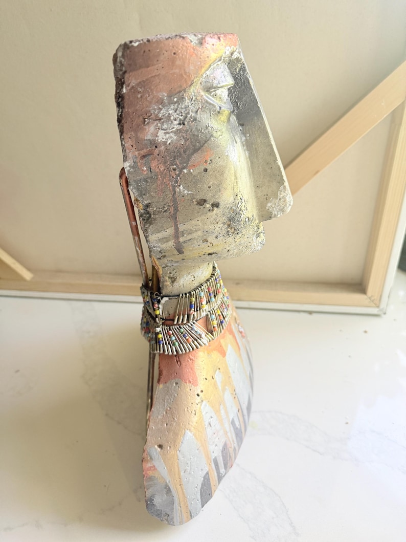 K&ouml;nnte beinhalten: Abstrakte Skulptur eines Kopfes und eines Torsos mit strukturierter, verwitterter Optik. Die Skulptur hat eine Farbpalette aus Silber, Orange und Wei&szlig; mit einer Perlenkette. Das Kunstwerk wird auf einer wei&szlig;en Oberfl&auml;che pr&auml;sentiert.