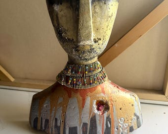 Può includere: Un busto in cemento raffigurante una testa e spalle umane con una finitura usurata e strutturata nei toni del giallo, nero e arancione. La figura indossa una collana a più strati composta da piccole perline colorate e spilli metallici.