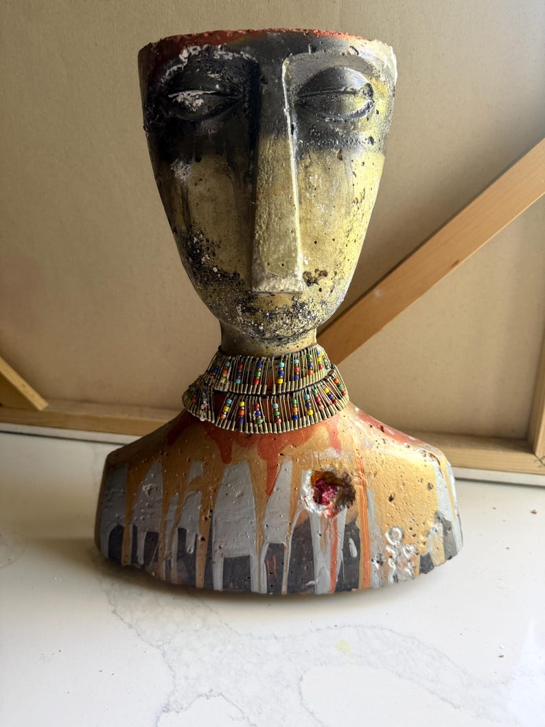 K&ouml;nnte beinhalten: Eine Betonb&uuml;ste eines menschlichen Kopfes mit Schultern in einer verwitterten, strukturierten Oberfl&auml;che in Gelb-, Schwarz- und Oranget&ouml;nen. Die Figur tr&auml;gt eine mehrlagige Halskette aus kleinen, bunten Perlen und metallischen Stiften.