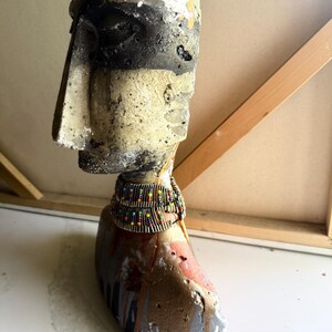K&ouml;nnte beinhalten: Eine verwitterte, abstrakte Skulptur eines Kopfes und Halses, mit einer neutralen Farbpalette aus Schwarz-, Grau- und Beiget&ouml;nen. Die Skulptur ist mit einem Perlenhalsband verziert und hat eine raue, strukturierte Oberfl&auml;che, die auf eine gealterte oder handgefertigte &Auml;sthetik hindeutet.