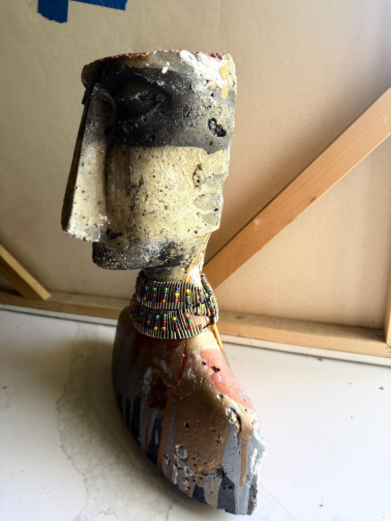 K&ouml;nnte beinhalten: Eine verwitterte, abstrakte Skulptur eines Kopfes und Halses, mit einer neutralen Farbpalette aus Schwarz-, Grau- und Beiget&ouml;nen. Die Skulptur ist mit einem Perlenhalsband verziert und hat eine raue, strukturierte Oberfl&auml;che, die auf eine gealterte oder handgefertigte &Auml;sthetik hindeutet.