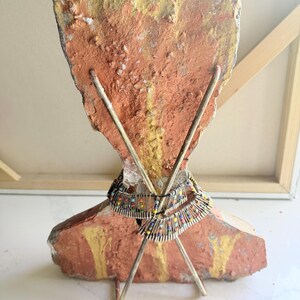 K&ouml;nnte beinhalten: Eine einzigartige Skulptur mit einem grob strukturierten, orange und gelb lackierten Steinsockel. Zwei Metallst&auml;be kreuzen sich davor und tragen eine Perlenkette mit farbenfrohen Akzenten. Das St&uuml;ck hat eine k&uuml;nstlerische, handwerkliche &Auml;sthetik.