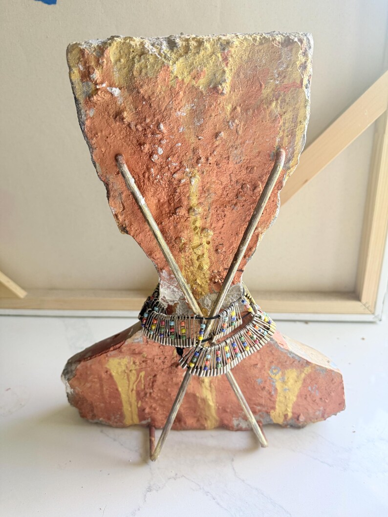 K&ouml;nnte beinhalten: Eine einzigartige Skulptur mit einem grob strukturierten, orange und gelb lackierten Steinsockel. Zwei Metallst&auml;be kreuzen sich davor und tragen eine Perlenkette mit farbenfrohen Akzenten. Das St&uuml;ck hat eine k&uuml;nstlerische, handwerkliche &Auml;sthetik.