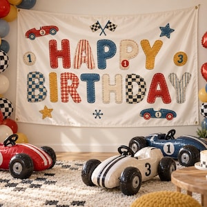 May include: A race car themed birthday party setup featuring a large white banner with the text HAPPY BIRTHDAY, colorful balloon arches, a red number 3 balloon, and three toy race cars in red, white, and blue with racing numbers on them.