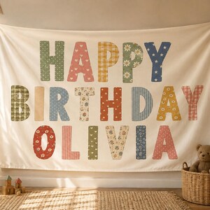 Puede incluir: Un tapiz de tela color crema con el texto HAPPY BIRTHDAY OLIVIA en letras grandes de estilo bloque. Cada letra muestra un patrón único, incluyendo diseños florales, de cuadros, de lunares y de estrellas en tonos tierra y pastel apagados.