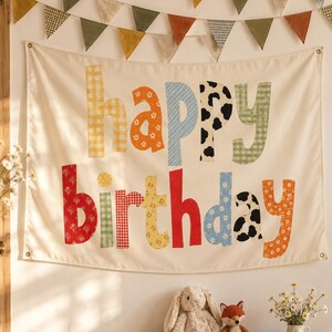 Może przedstawiać: Kremowy baner z tkaniny z napisem "happy birthday" wykonanym z dużych, wielokolorowych i wzorzystych liter. Litery zawierają r&oacute;żne nadruki, takie jak kratka vichy, kwiaty i krowie łaty. Powyżej zawieszona jest girlanda z tr&oacute;jkątnych proporczyk&oacute;w.