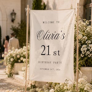 Könnte beinhalten: Ein rechteckiges, cremefarbenes Stoffbanner, das zwischen zwei Holzstangen gespannt ist. Das Banner trägt den schwarzen Aufdruck: WELCOME TO Olivia's 21 st BIRTHDAY PARTY SEPTEMBER 26TH, 2026. Der Stoff ist mit kleinen weißen Bändern befestigt.